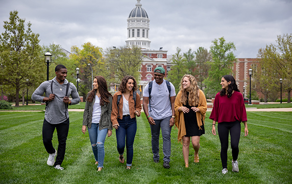 Photo of Jesse Hall with six students walking in grass on a cloudy day.